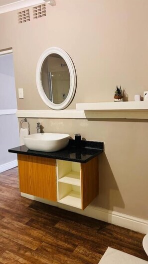 Un baño con lavamanos blanco, encimera negra, armario de madera y espejo redondo.
