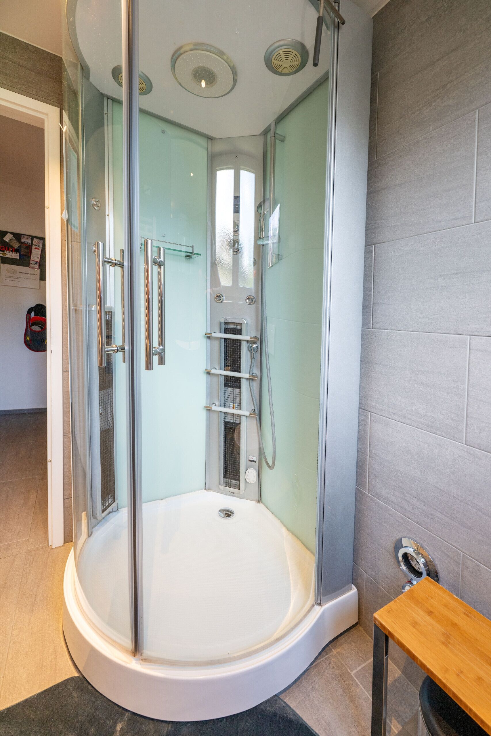 A glass-enclosed shower stall with a white bathtub, a wooden bench, and a tiled floor.