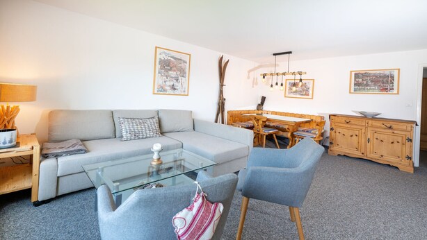 Ein Wohnzimmer mit Sofa, Sesseln, einem Glastisch, einem Esstisch mit Stühlen, Holzsäcken und an der Wand befestigten Bildern.