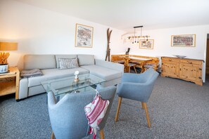 A living room with a sofa, chairs, a glass coffee table, a dining table with chairs, wooden cabinets, and wall-mounted pictures.