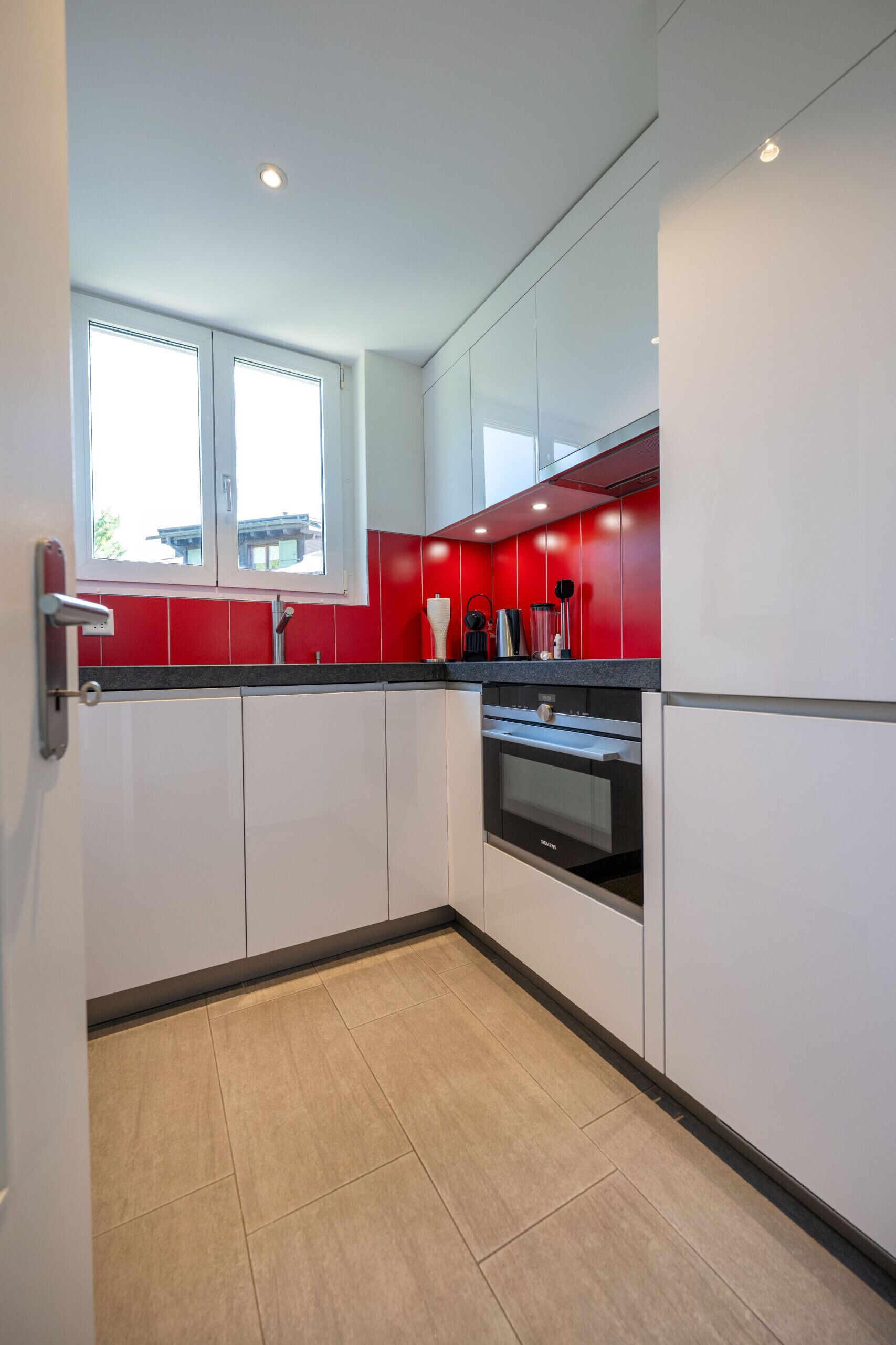 Une cuisine avec des meubles blancs, un crédence rouge, un four noir et une fenêtre au-dessus de l’évier.