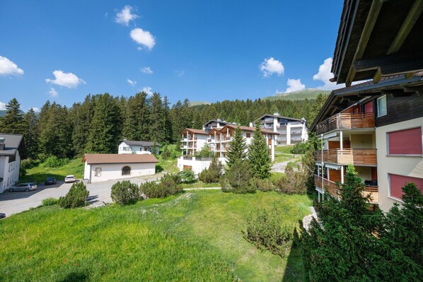 A residential area with multi-story buildings, a grassy foreground, and a forested hillside in the background.