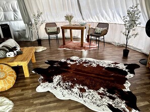 Una habitación con toldo, una mesa de madera, dos sillas, un tapiz de cuero de vaca y una planta en maceta.