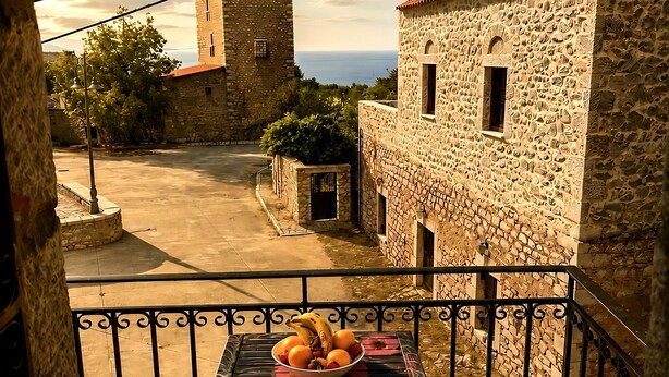 A balcony with a table and chairs, a bowl of fruit, and a view of a stone building and the sea. - Diak’s Tower | Sunset Sea View in Mani (Charia)