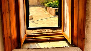 A room with a wooden-framed window overlooking a courtyard with a stone building and greenery. - Diak’s Tower | Sunset Sea View in Mani (Charia)