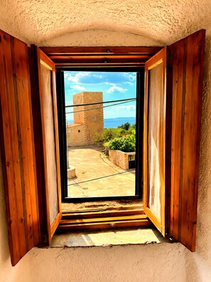 Ein Zimmer mit einem raumhohen, hölzernen Fenster, das auf einen Innenhof mit einem Steinhaus und viel Grün blickt.