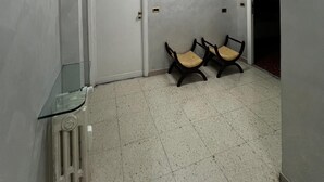 A hallway with a white door, two wooden chairs, a glass table, and a radiator. - Accommodation in Appia Nuova (Roma)