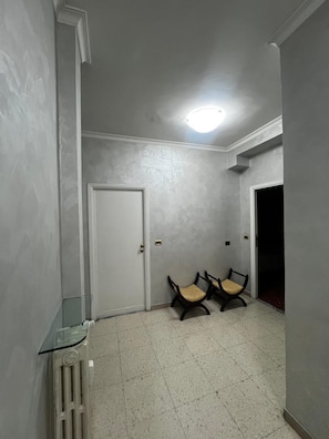 A hallway with a white door, two wooden chairs, a glass table, and a radiator. - Accommodation in Appia Nuova (Roma)