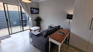 A modern living room with a sofa, a small table, a TV, and a sliding glass door leading to an outdoor area. - Saxon Guest Cottage Suite (Cape Town)