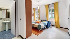 A bedroom with a bed, a bedside table, a window with curtains, a radiator, and a bathroom with a sink and mirror. - Varazze Mare Apartment with Two Bathrooms and Courtyard (Varazze)