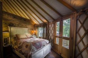 A cozy bedroom with a wooden ceiling, a bed with a patterned bedspread, a nightstand with a lamp, and a window with a lattice design. - Heavenly Yurt with Modern Interior in Topton, North Carolina (Topton)