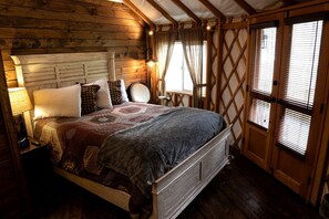A wooden cabin-style bedroom with a bed, nightstand, lamp, and a window with blinds. - Heavenly Yurt with Modern Interior in Topton, North Carolina (Topton)