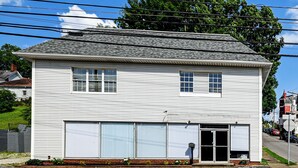 A two-story white building with a garage door, a small porch with plants, and a street view with cars and a sidewalk.
