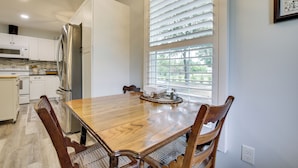 Une cuisine moderne avec des meubles blancs, un réfrigérateur en acier inoxydable, une table à manger en bois et une fenêtre avec des stores.
