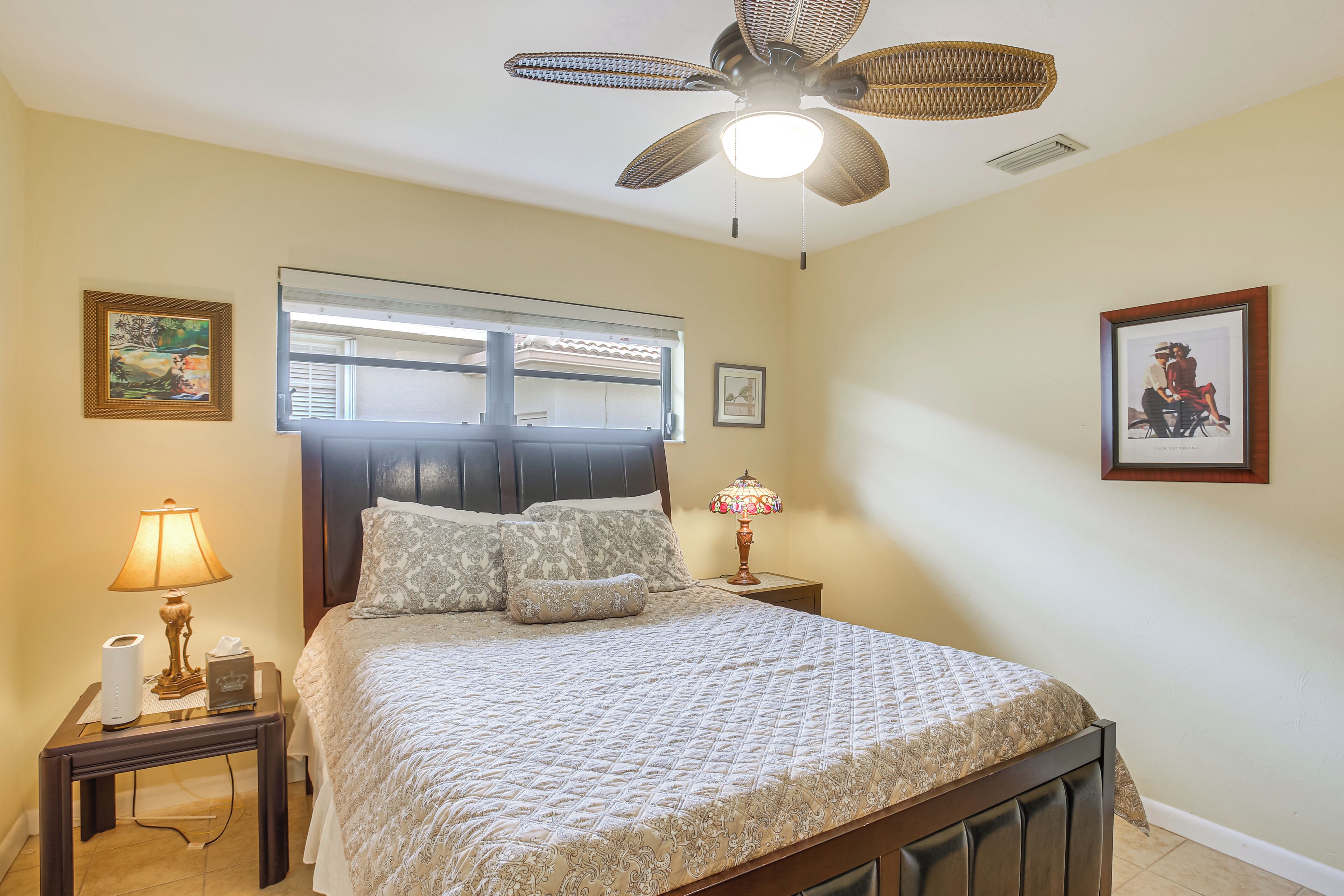 Un dormitorio con cama, mesita de noche, lámpara, ventilador de techo y dos cuadros en la pared.