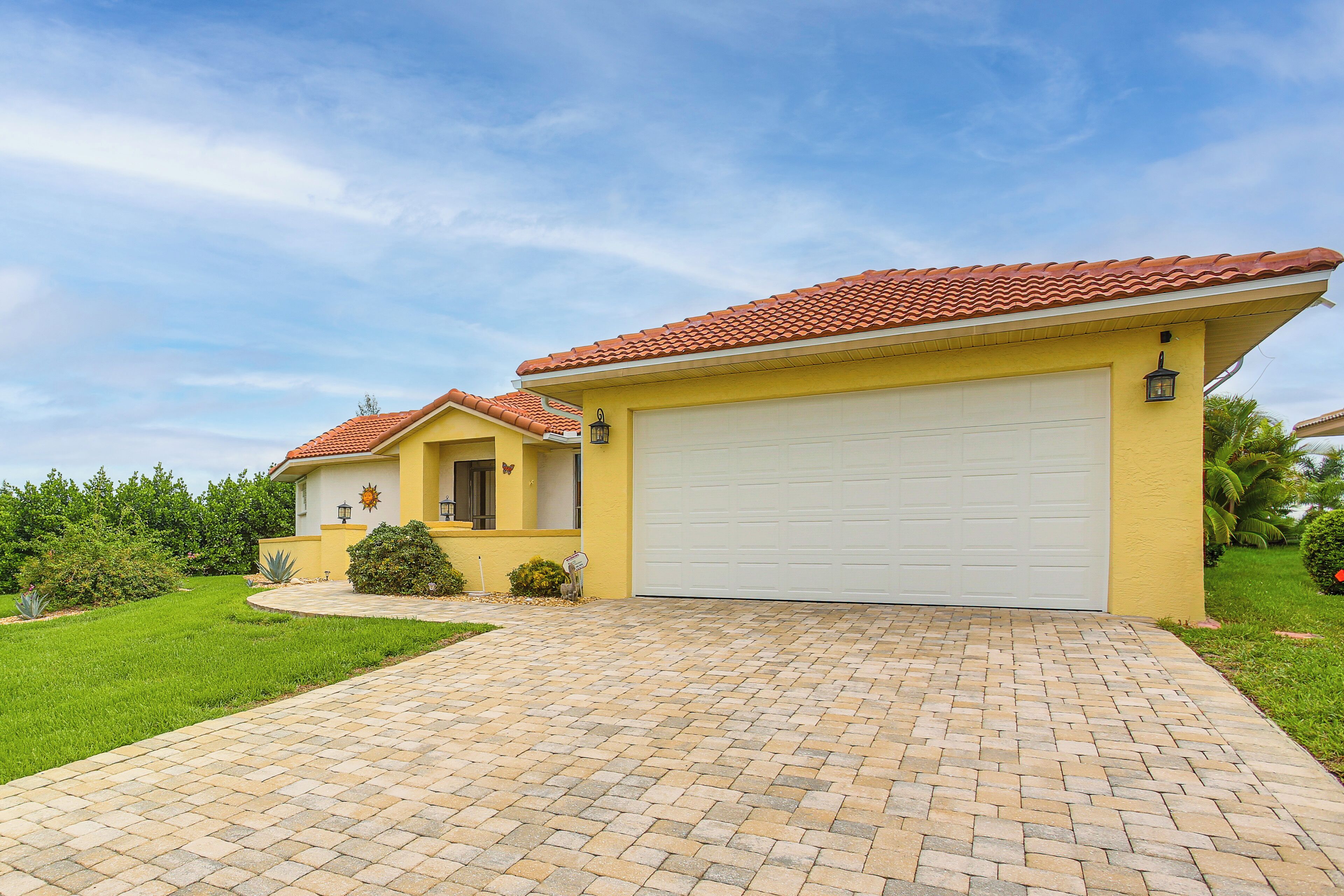 A residential house with a red-tiled roof, a yellow facade, and a paved driveway leading to a white garage door.