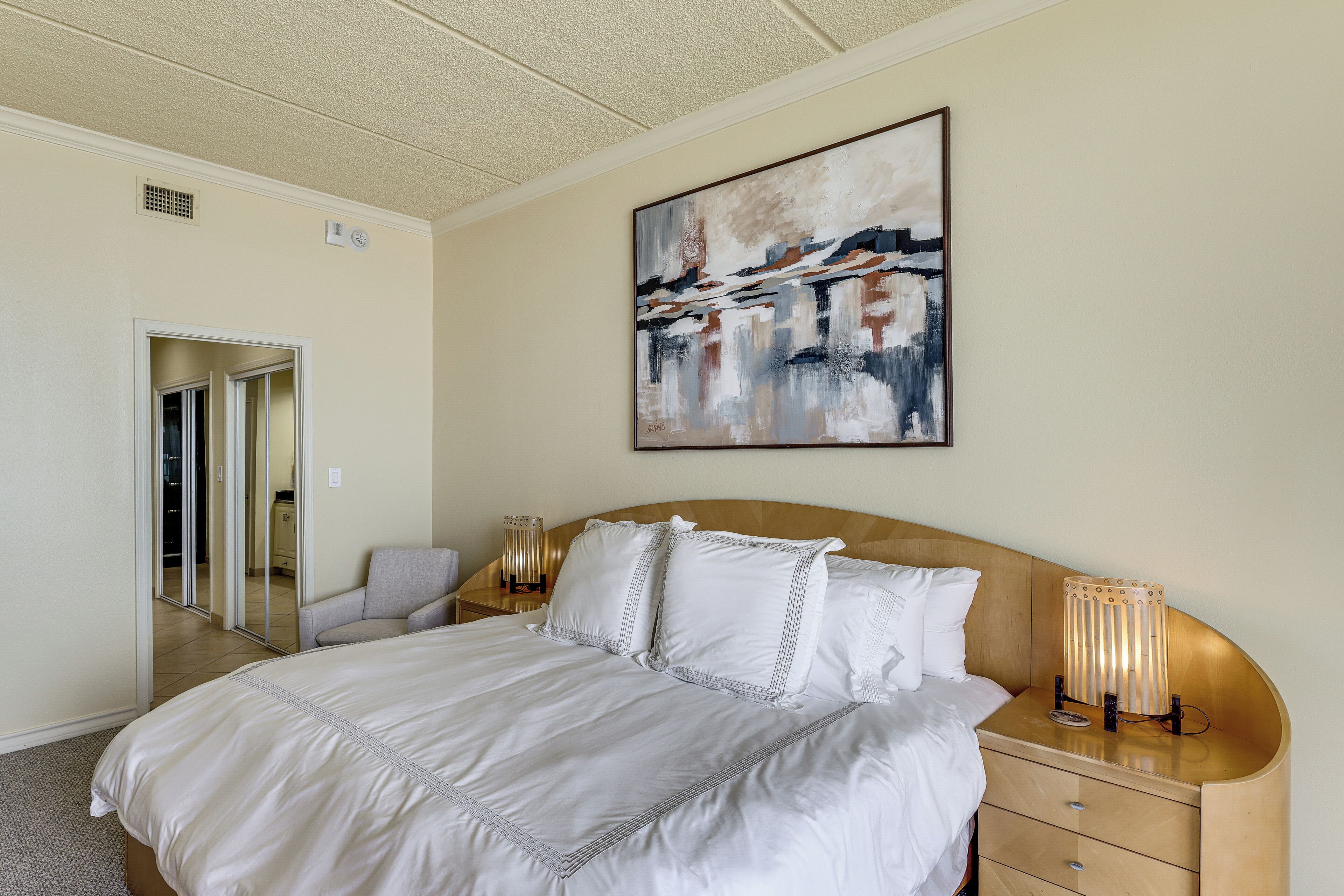A hotel room with a large bed, a bedside table with a lamp, a painting on the wall, and a door leading to another room.