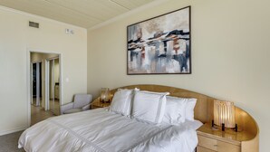 A hotel room with a large bed, a bedside table with a lamp, a painting on the wall, and a door leading to another room. - 3BR Beachfront PH Suite: Suntide III - Unit 1203 (South Padre Island)