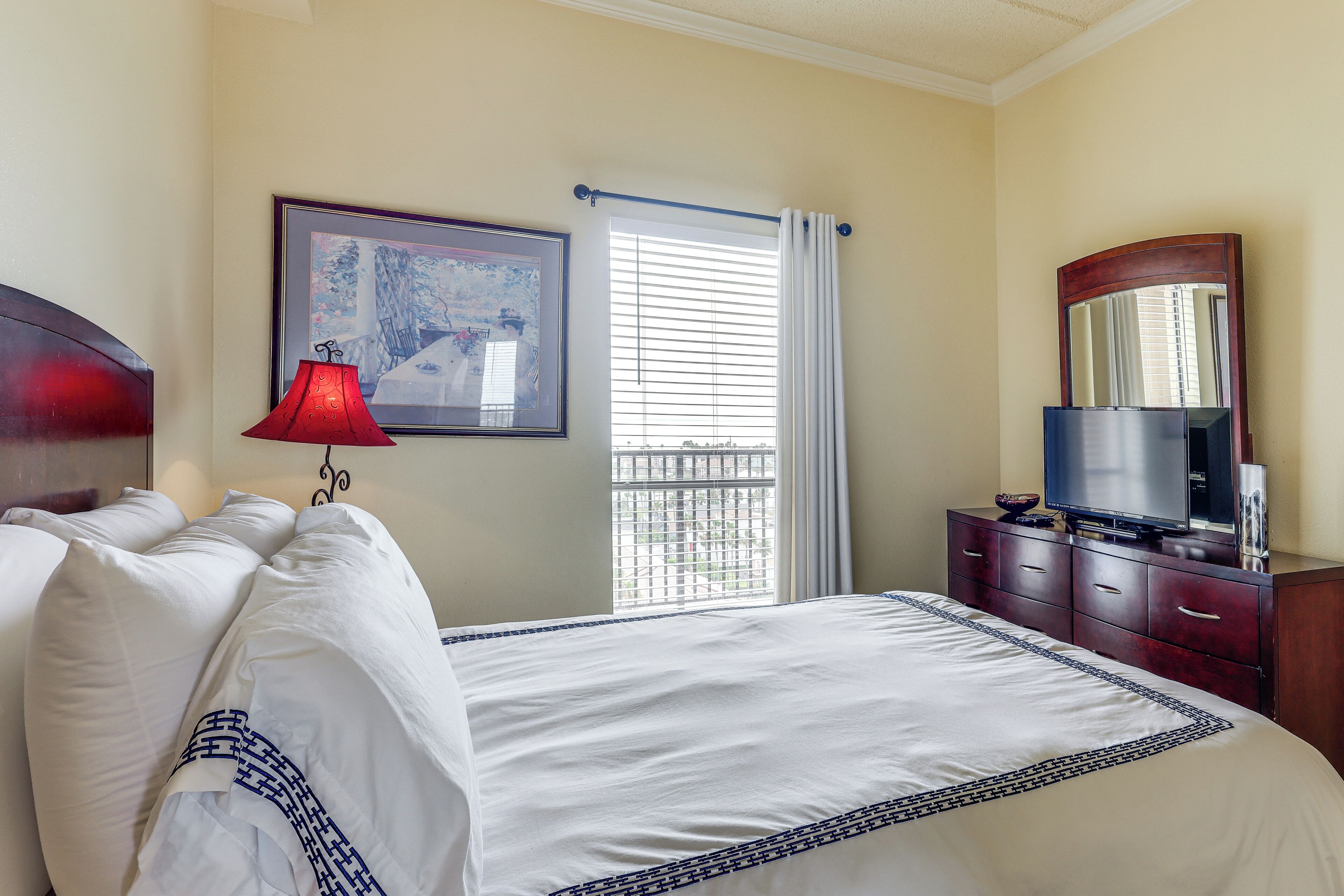 Una habitación de hotel con cama, televisión, cómoda, ventana con persianas y un cuadro en la pared.