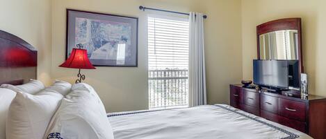 Una habitación de hotel con cama, televisión, cómoda, ventana con persianas y un cuadro en la pared.