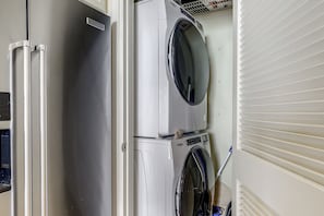 Ein kleiner Waschraum mit einer übereinander gestapelten, weißen Waschmaschine und Trockner, einem Edelstahlkühlschrank und einem an der Wand angelehnten Mop.
