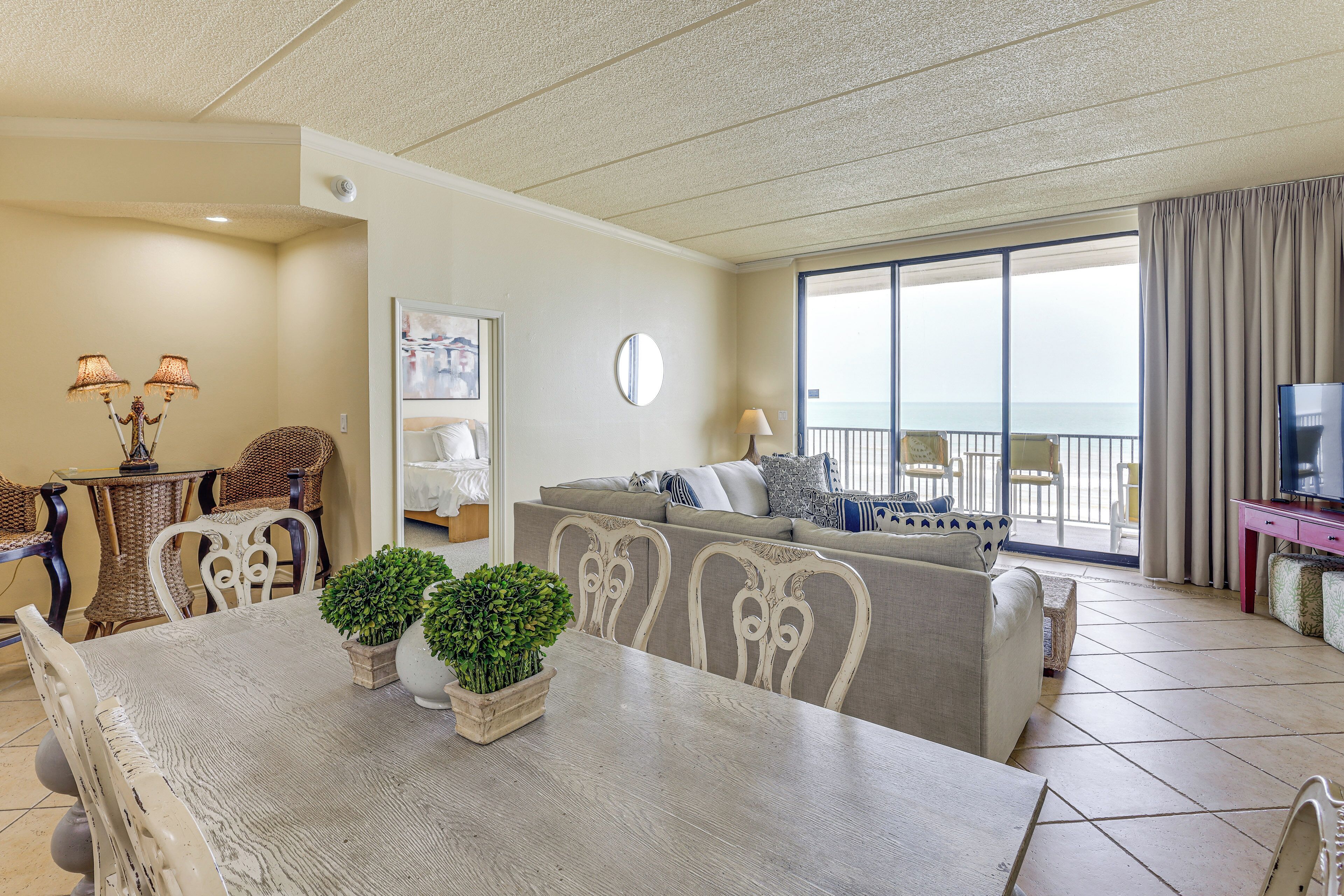 A living room with a dining table, chairs, a sofa, a TV, and a balcony with a view.