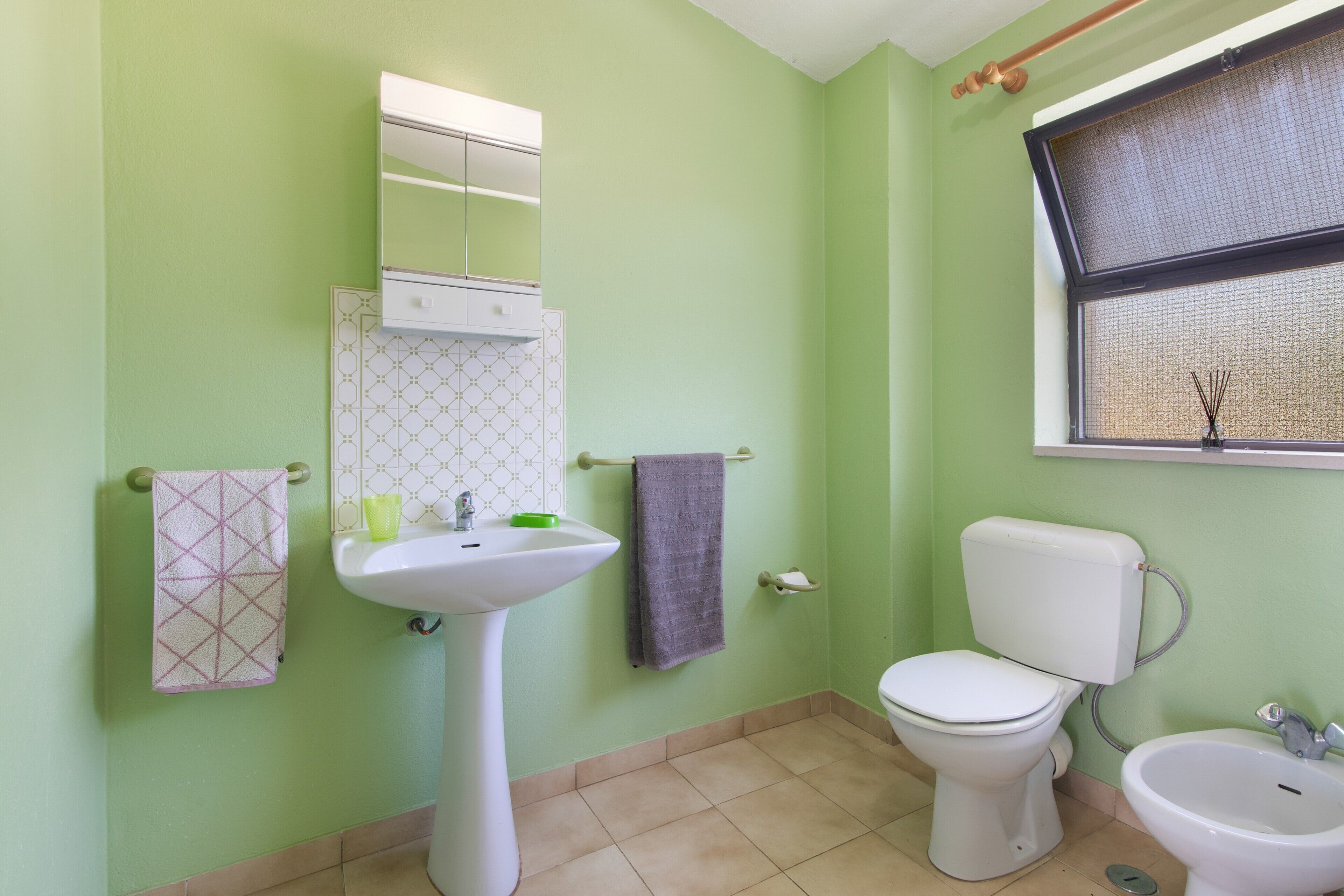 A bathroom with a toilet, bidet, sink, and a window with a blind.