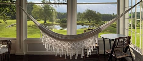A room with a hammock, a round table, and chairs, overlooking a green landscape with trees and a body of water.
