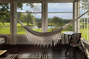 A room with a hammock, a round table, and chairs, overlooking a green landscape with trees and a body of water.