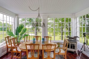A dining room with a round table, wooden chairs, a chandelier, and large windows with a view of greenery.