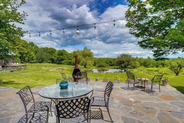 A patio with a glass table and metal chairs, surrounded by stone flooring, with a view of a pond and trees.