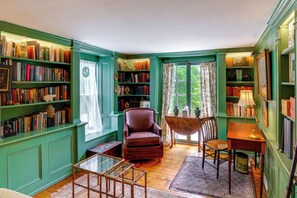 A room with green walls, wooden flooring, a leather armchair, a wooden table, a glass coffee table, and a bookshelf filled with books.
