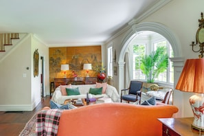 A spacious living room with a large sofa, a coffee table, and a fireplace. There is a staircase leading to another level, and a large window with a view of greenery outside.