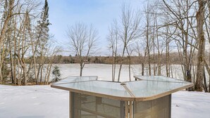 Un espacio exterior cubierto de nieve con una estructura metálica, posiblemente un refugio, rodeado de árboles sin hojas.