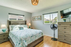 A bedroom with a bed, bedside lamps, a standing fan, a TV, and a window with blinds.