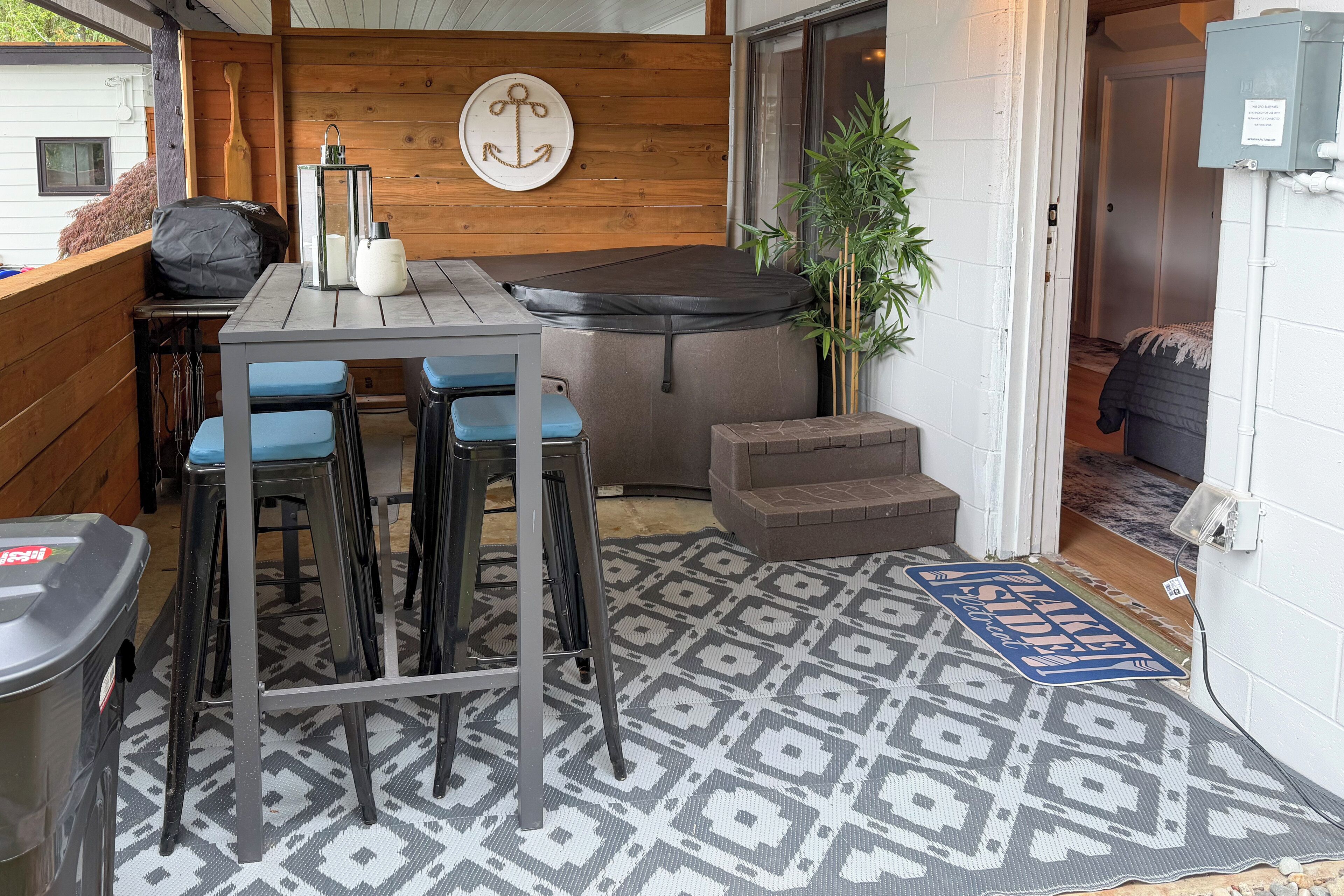 A compact outdoor seating area with a table and chairs, a small step stool, a decorative anchor wall hanging, and a bamboo plant.