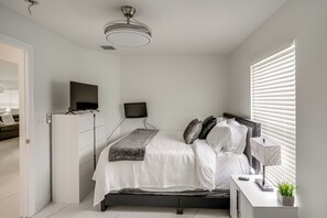 Ein Schlafzimmer mit Bett, Fernseher, Schreibtisch mit Lampe und einem Fenster mit Jalousien.
