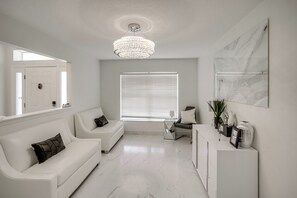 Ein modernes Wohnzimmer mit einem weißen Sofa, einem Couchtisch, einer Kristallleuchtre und einem großen Fenster mit Jalousien.