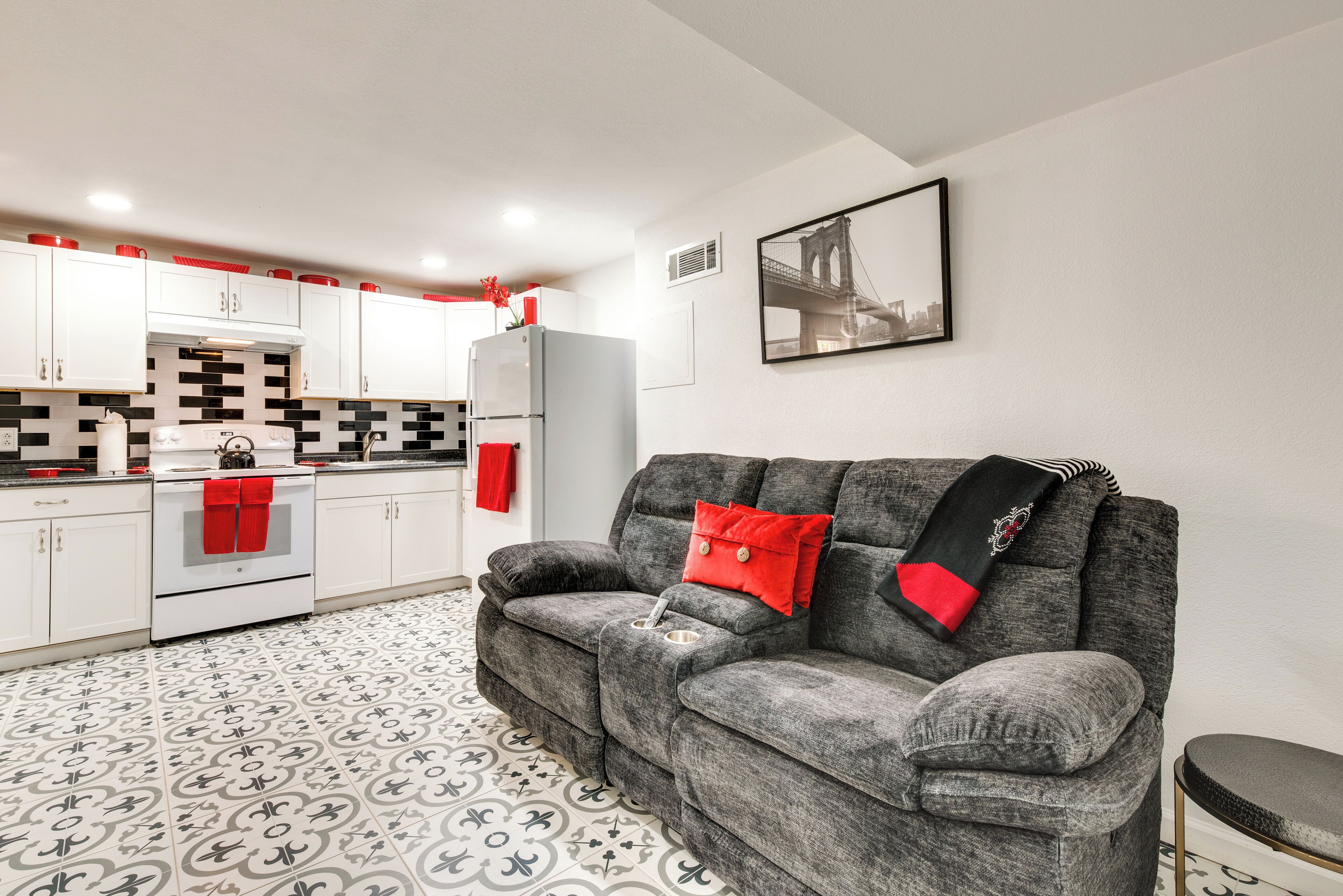 A compact living space with a kitchenette, grey sofa with red cushions, patterned floor tiles, and a framed picture on the wall.