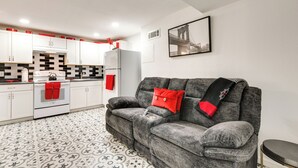 A compact living space with a kitchenette, grey sofa with red cushions, patterned floor tiles, and a framed picture on the wall.