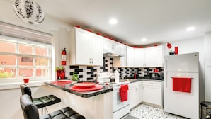 Una cocina moderna con armarios blancos, salpicadero de baldosas blanco y negro, un refrigerador con un toalla roja y una ventana con persianas.