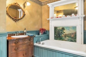 A bathroom with a wooden vanity, a white bathtub, a large mirror, and a fireplace with a painting. - Hot Tub & Large Yard: Elegant Deansboro Getaway (Deansboro)