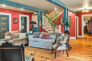 A spacious living room with a staircase, wooden flooring, and a mix of furniture including a patterned sofa and a classic armchair. - Hot Tub & Large Yard: Elegant Deansboro Getaway (Deansboro)