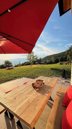 Une table de pique-nique en bois, avec un saladier rempli de collations, un parasol rouge et une vue pittoresque sur un champ verdoyant et des montagnes.