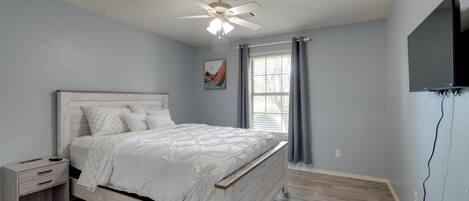 A bedroom with a bed, a nightstand, a ceiling fan, a window with curtains, and a wall-mounted TV.