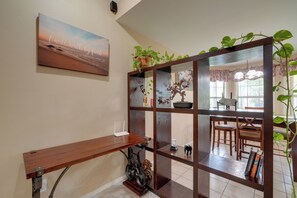 Una habitación con escritorio de madera, estantería, planta en maceta y zona de comedor con sillas y mesa.