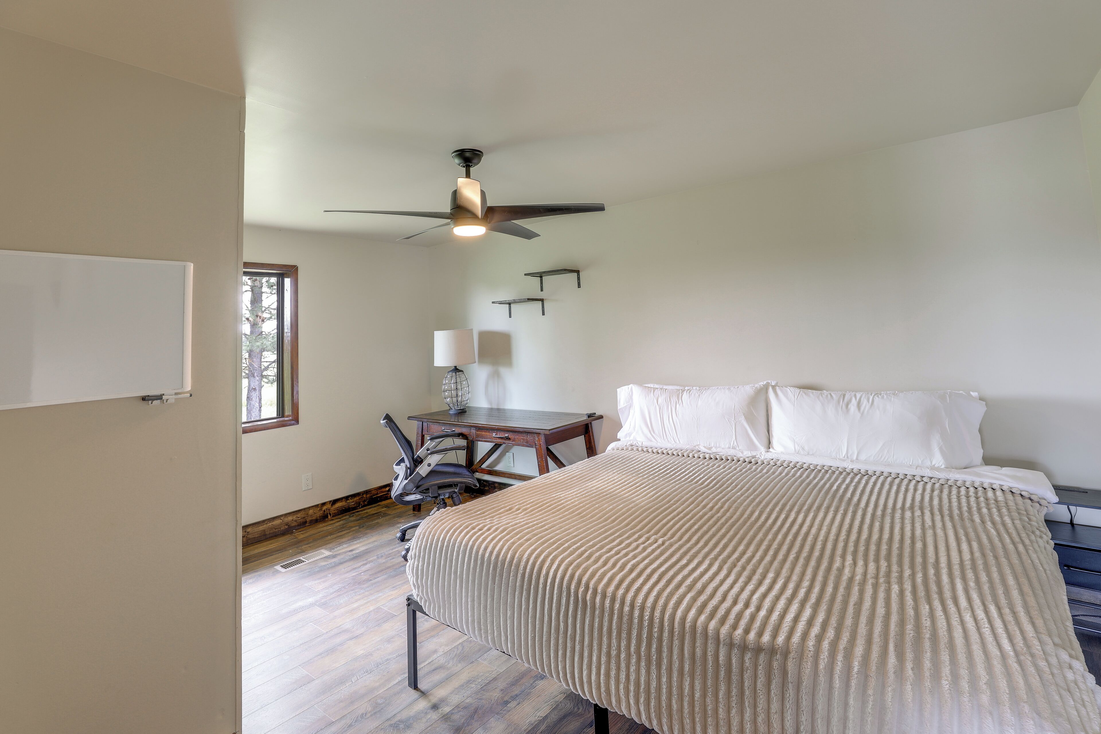 A bedroom with a bed, a desk, a chair, a ceiling fan, and a window.