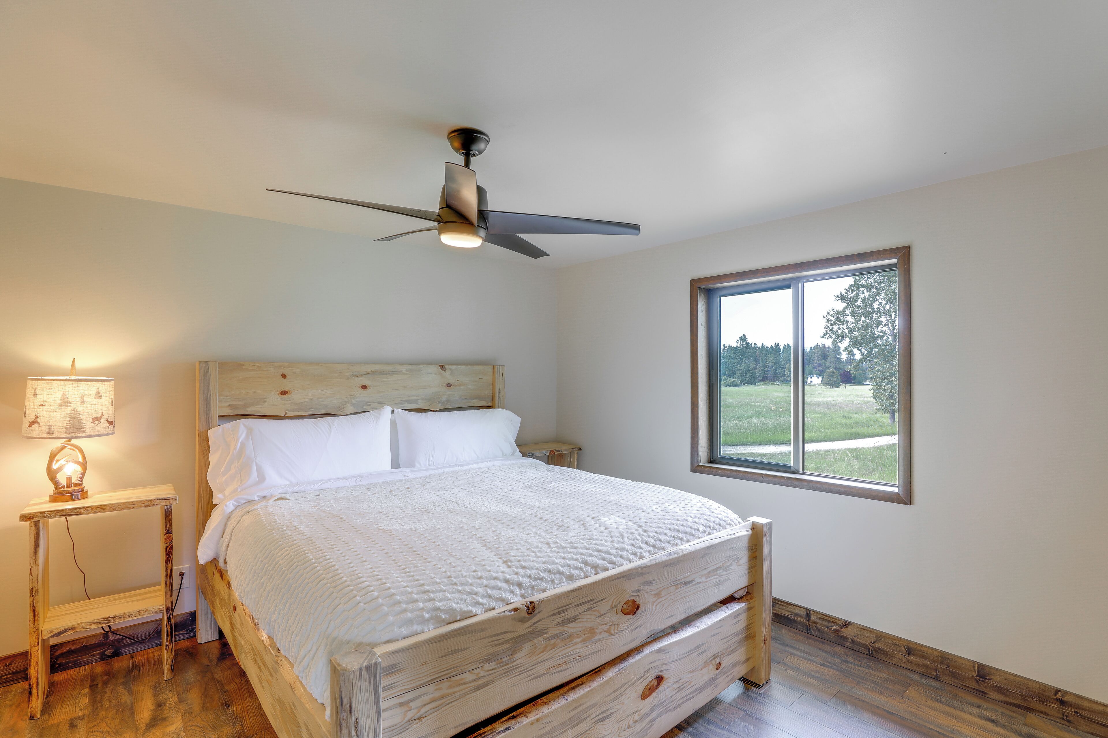 A bedroom with a bed, a ceiling fan, a nightstand with a lamp, and a window with a view of the outdoors.