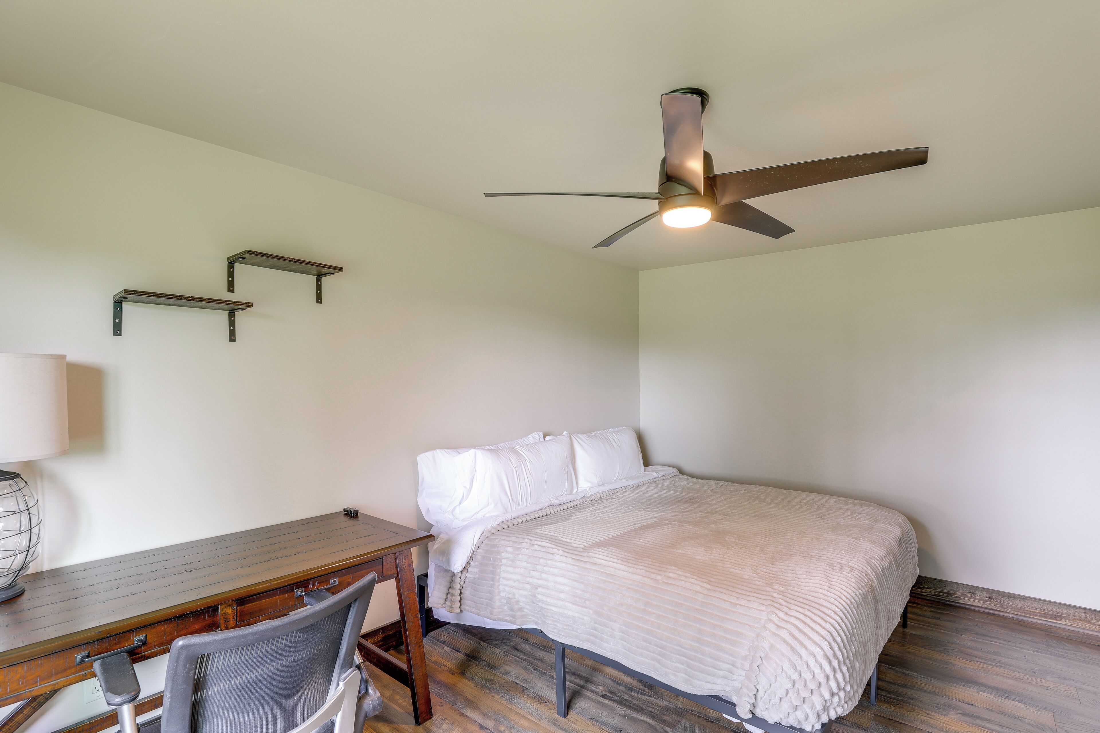 A bedroom with a bed, a desk, a chair, a ceiling fan, and wall-mounted shelves.