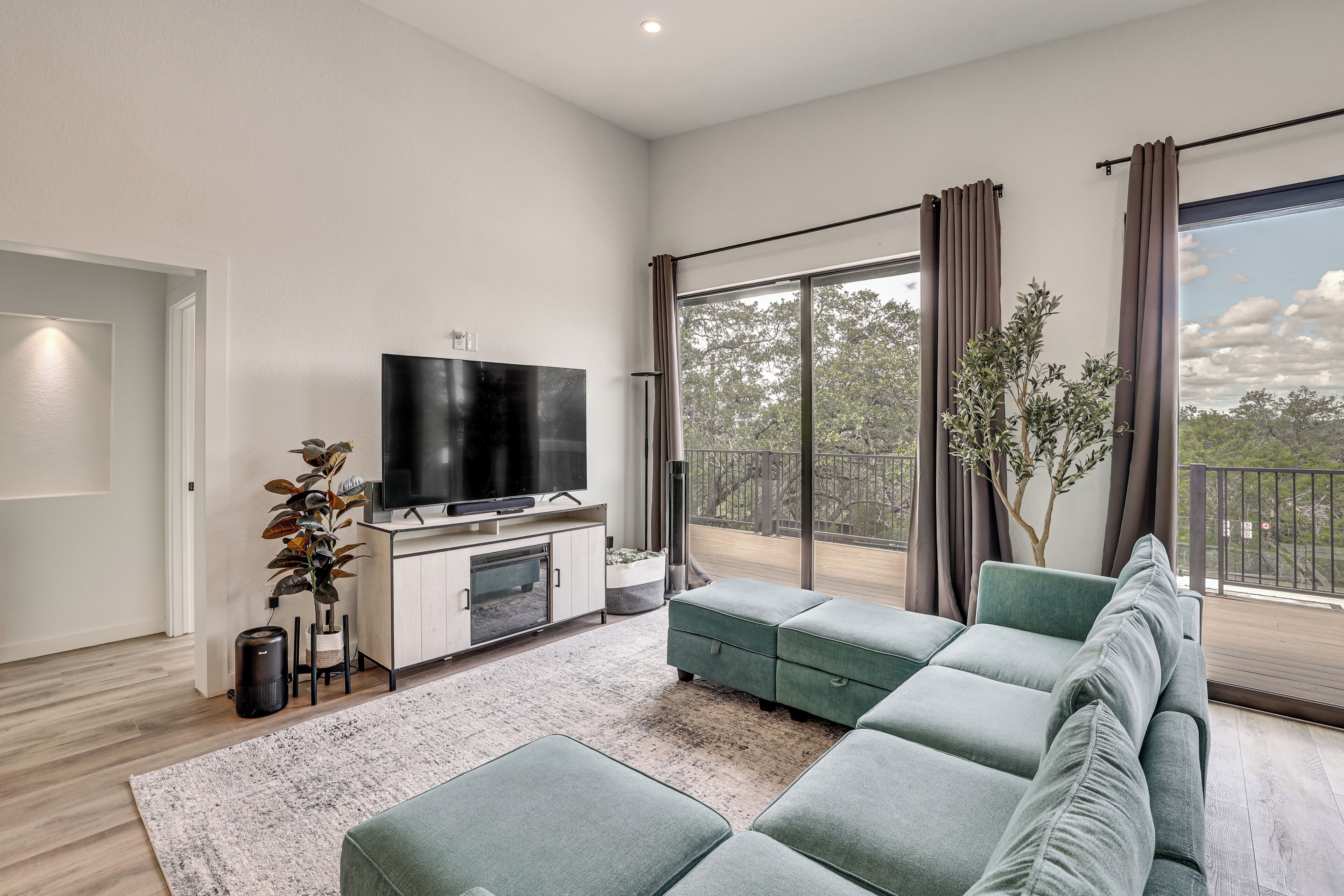 A modern living room with a sectional sofa, a flat-screen TV, a large window with curtains, and a potted plant.
